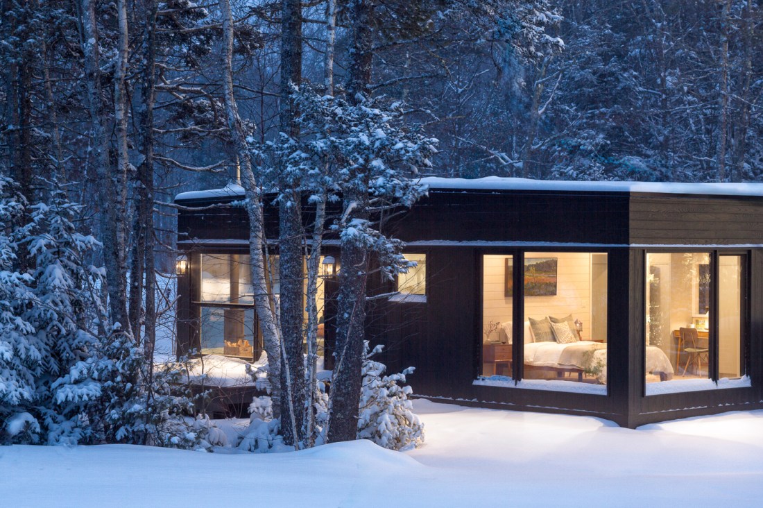 A modern black cabin with a flat roof nestled in a snow-covered pain forest at dusk. There is warm lighting visible inside through large windows. Outside, fresh snow covers the ground and tree branches. 