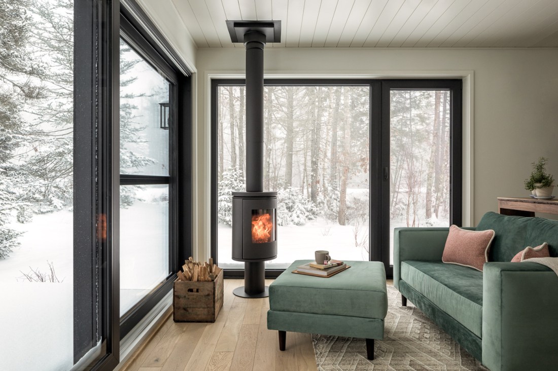 Minimalist living room corner with floor-to-ceiling windows overlooking a snowy winter forest. There is a modern wood-burning stove and a sage green sofa and ottoman. 