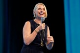 Elizabeth Gilbert, a white woman wearing a blue blouse, smiles while speaking into a microphone.