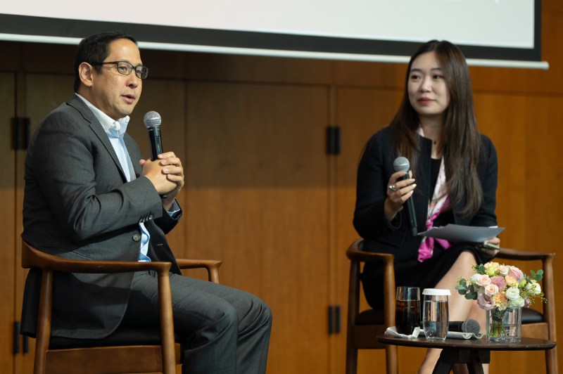 Two panelists sitting on stage in wooden chairs holding microphones. One panelist is actively speaking, the other is listening. Both are wearing business casual attire. 