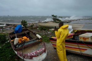 A person in a yellow waterproof suit standing next to two boats pulled ashore on a beach. The waves hitting the shore are large and rough and the sky is dark and grey as Hurricane Melissa approaches.