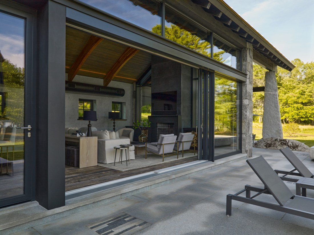 A modern stone house with floor to ceiling sliding glass doors fully opened to reveal a minimalist kitchen and dining area with exposed ceiling beams. There is a concrete patio with contemporary lounge chairs and the house is surrounded by green trees. 