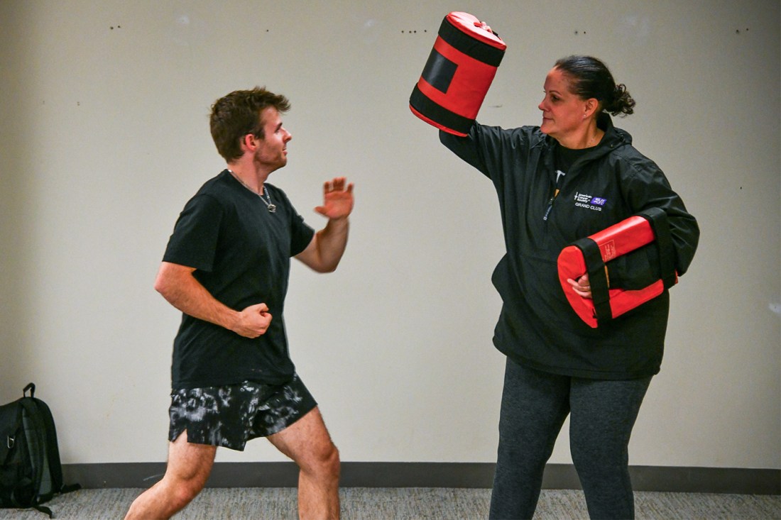 A man in a black t-shirt and patterned shorts practices defensive movements while an instructor in a black jacket and leggings holds a red and black strike pad overhead.