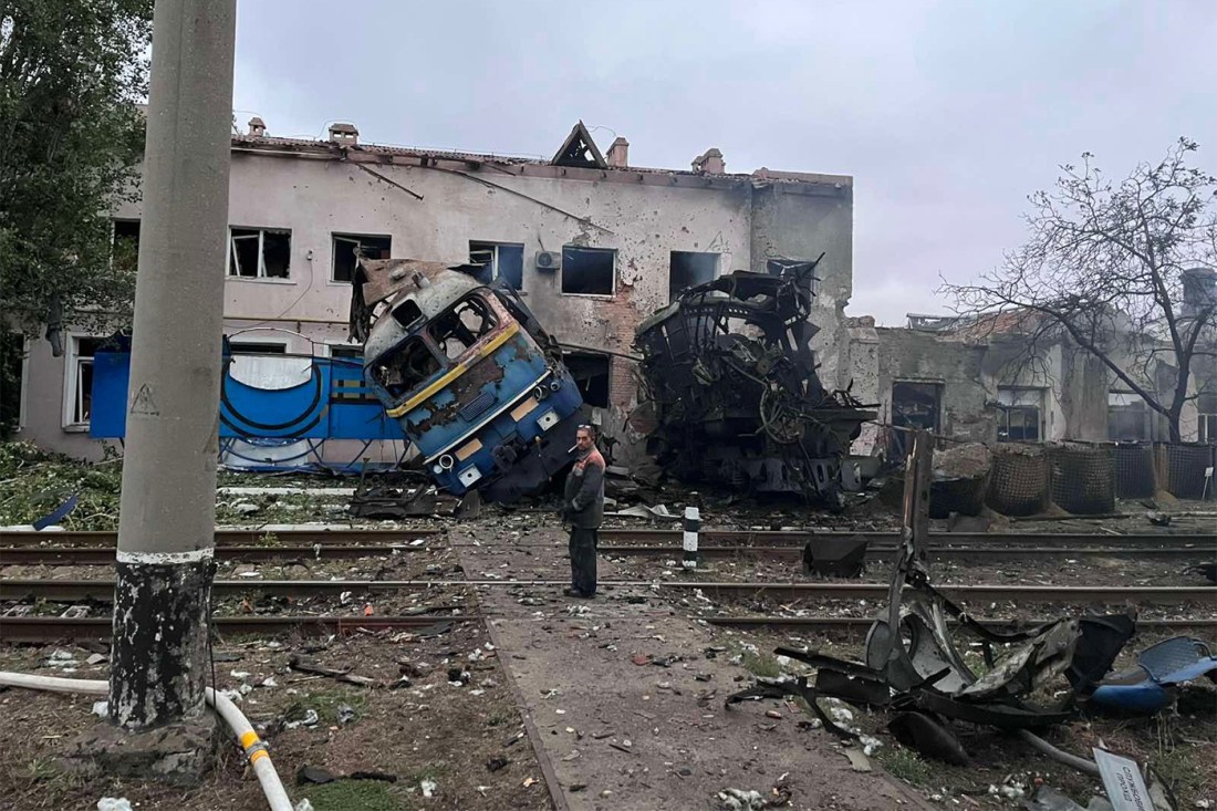 A railway worker stands in front of a destroyed train locomotive under a gray sky.