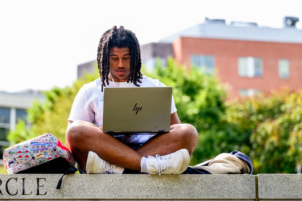 Trystan DaSilva sitting cross legged on a wall with his laptop balanced in his lap.