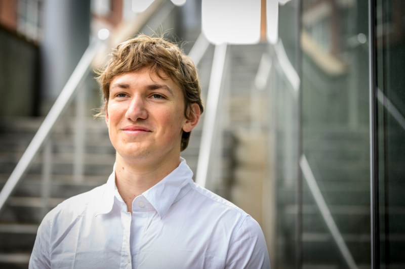 Pedro Graziosi, a biology student, stands outside wearing a white shirt, smiling slightly with a staircase and glass railings behind him.