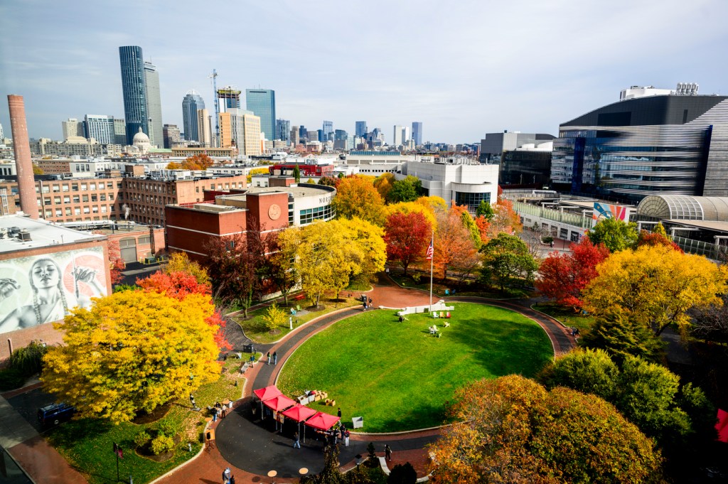 Aerial view of Centennial Common showing vibrant fall foliage in yellow, orange, and red surrounding a circular green lawn, with Boston's skyline visible in the background.